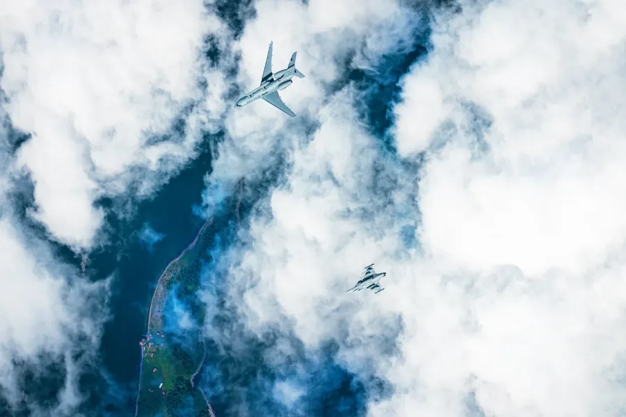 Two jets fly over a narrow green island surrounded by a blue sea, viewed through scattered white clouds, creating a dynamic aerial scene.