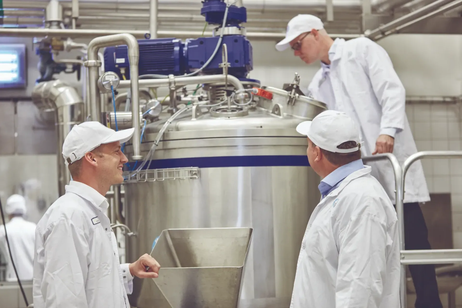 Three workers in white uniforms and caps inspect a large stainless steel industrial tank in a factory. They appear focused and engaged in discussion.