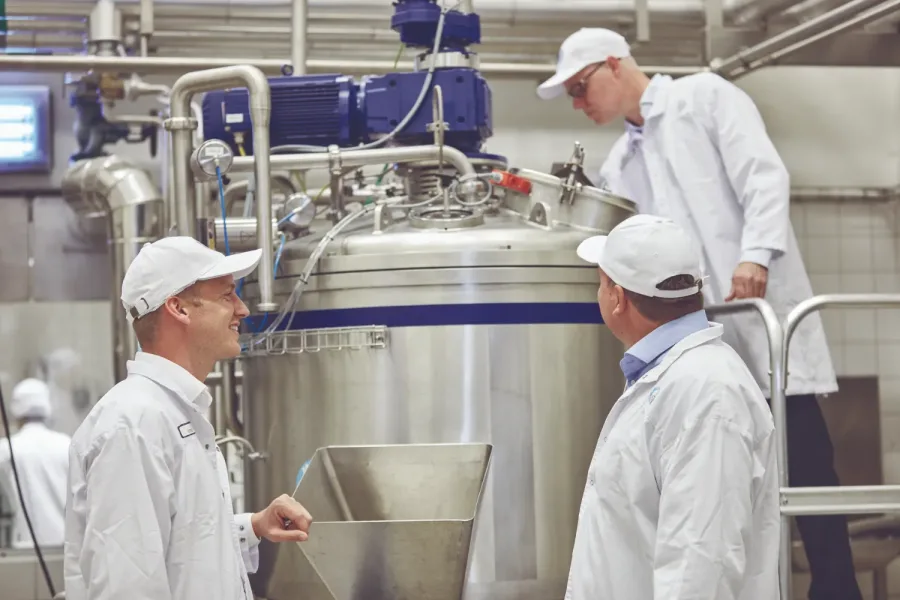 Three workers in white uniforms and caps inspect a large stainless steel industrial tank in a factory. They appear focused and engaged in discussion.