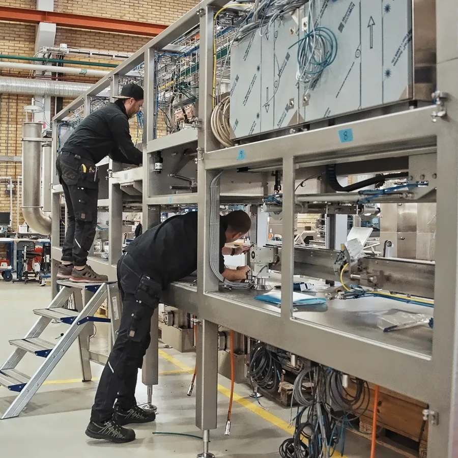 Two technicians in a factory setting work on a large industrial machine. One stands on a ladder, both focused and engaged in complex assembly.
