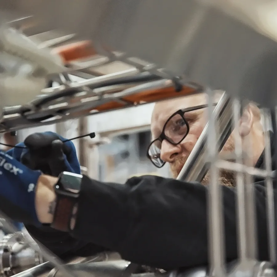 A person with glasses intently works on machinery, using a tool. They wear blue gloves and a dark sweater, conveying focus and technical skill.