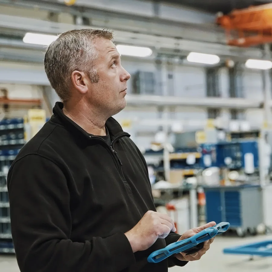 Man in a black jacket, holding a blue tablet, looks up thoughtfully in an industrial workshop setting with shelves and machinery in the background.