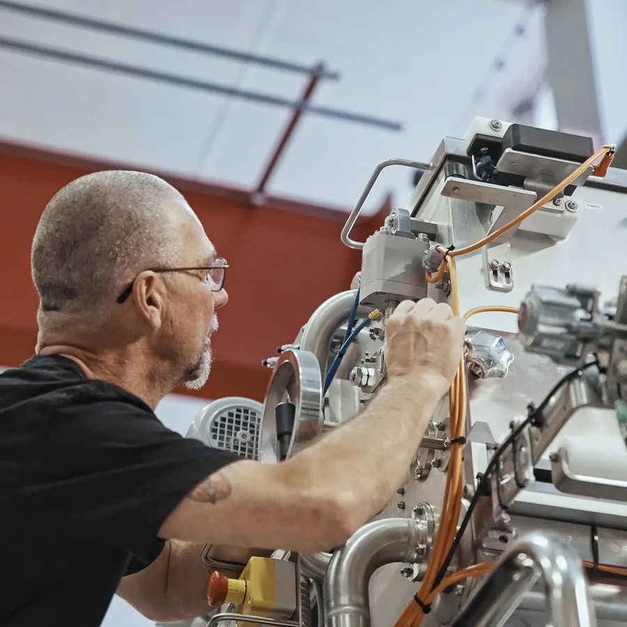 A technician with glasses and a beard works on a complex machine with orange cables. The setting is industrial, conveying focus and precision.