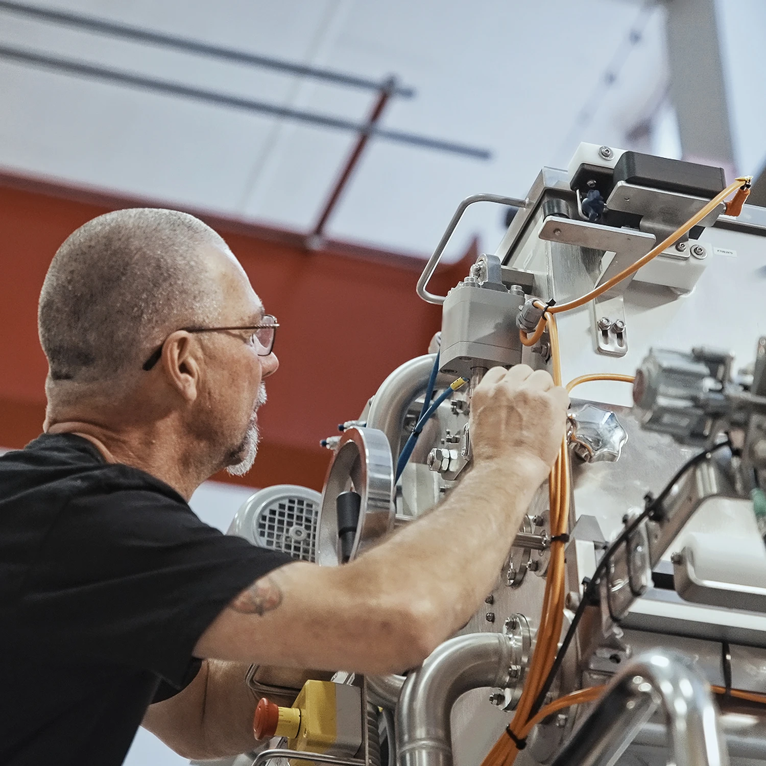 A technician with glasses and a beard works on a complex machine with orange cables. The setting is industrial, conveying focus and precision.