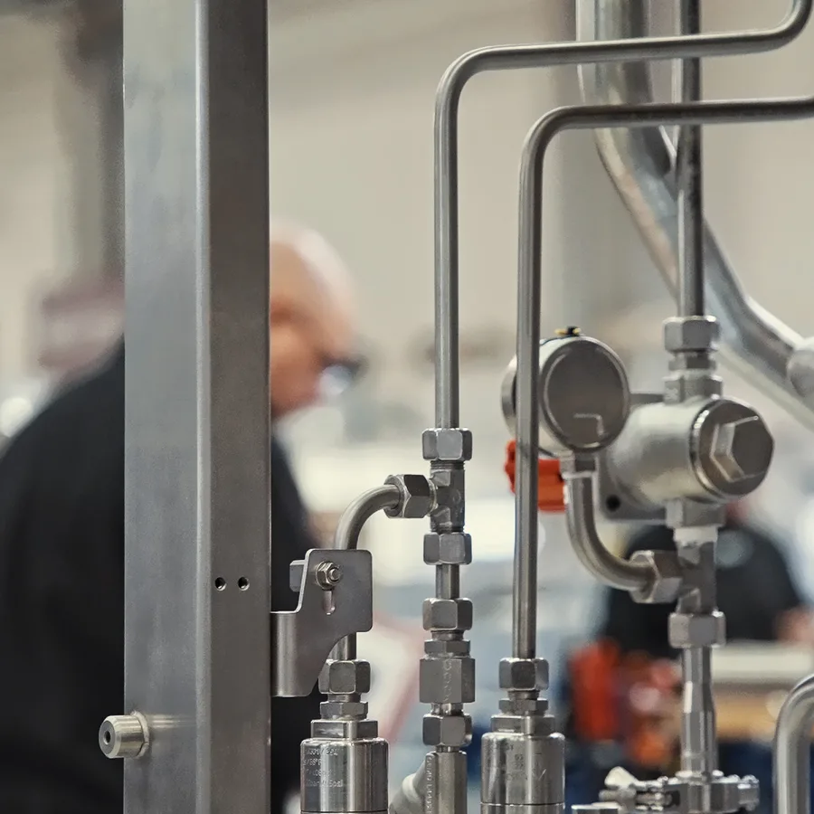 Close-up of industrial metal pipes and gauges with a blurred figure in the background, suggesting a manufacturing or workshop setting.
