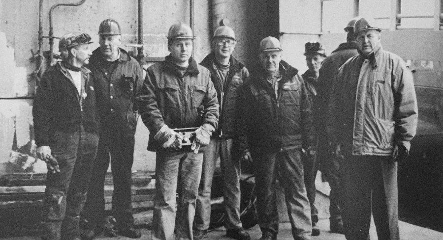 A group of eight men wearing work jackets and helmets stands together in an industrial setting. The scene conveys a sense of camaraderie and determination.