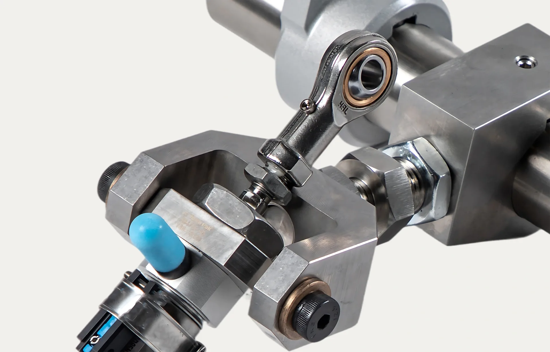 Close-up of a metallic mechanical component with bolts and bearings. A blue-tipped rubber part adds contrast. The polished metal conveys precision.