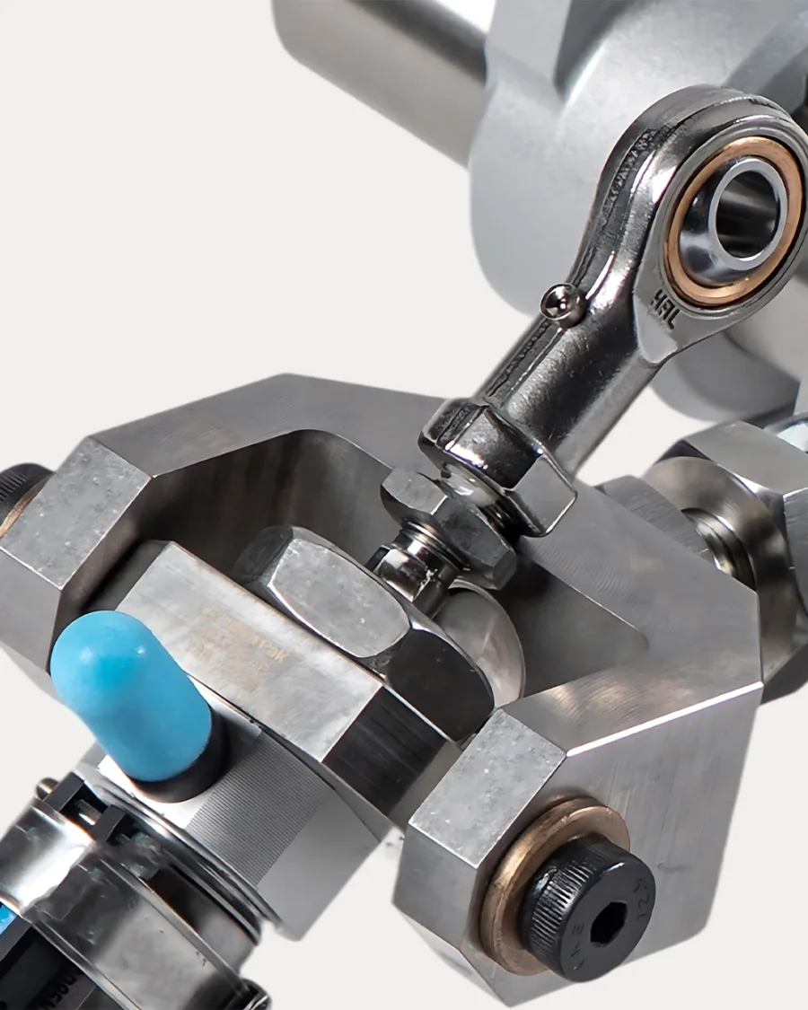 Close-up of a metallic mechanical component with bolts and bearings. A blue-tipped rubber part adds contrast. The polished metal conveys precision.