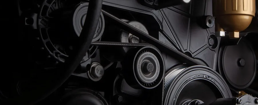 Close-up of a complex engine with black belts, gears, and metal components. A brass-colored filter stands out, conveying a technical and industrial feel.