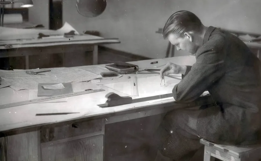 A man in a suit focuses intently on architectural drawings at a drafting table, surrounded by rulers and papers, in a vintage setting conveying concentration.