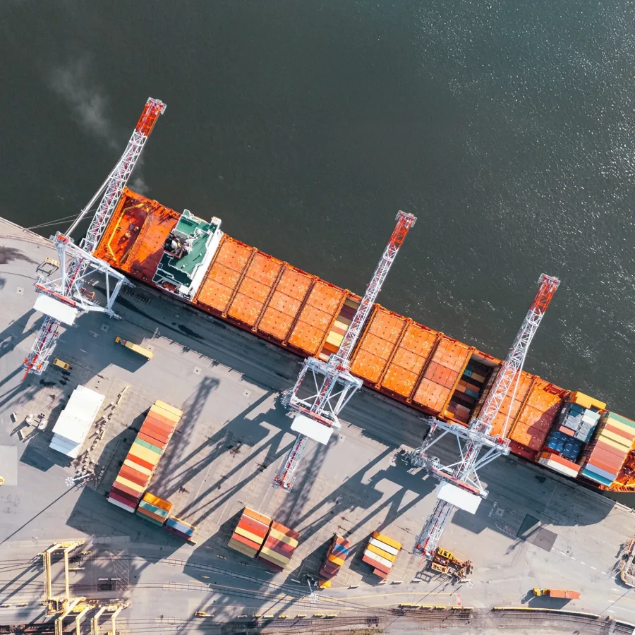 Aerial view of a cargo ship docked at a port, with orange containers on deck. Three large cranes and stacks of colorful containers are visible onshore.