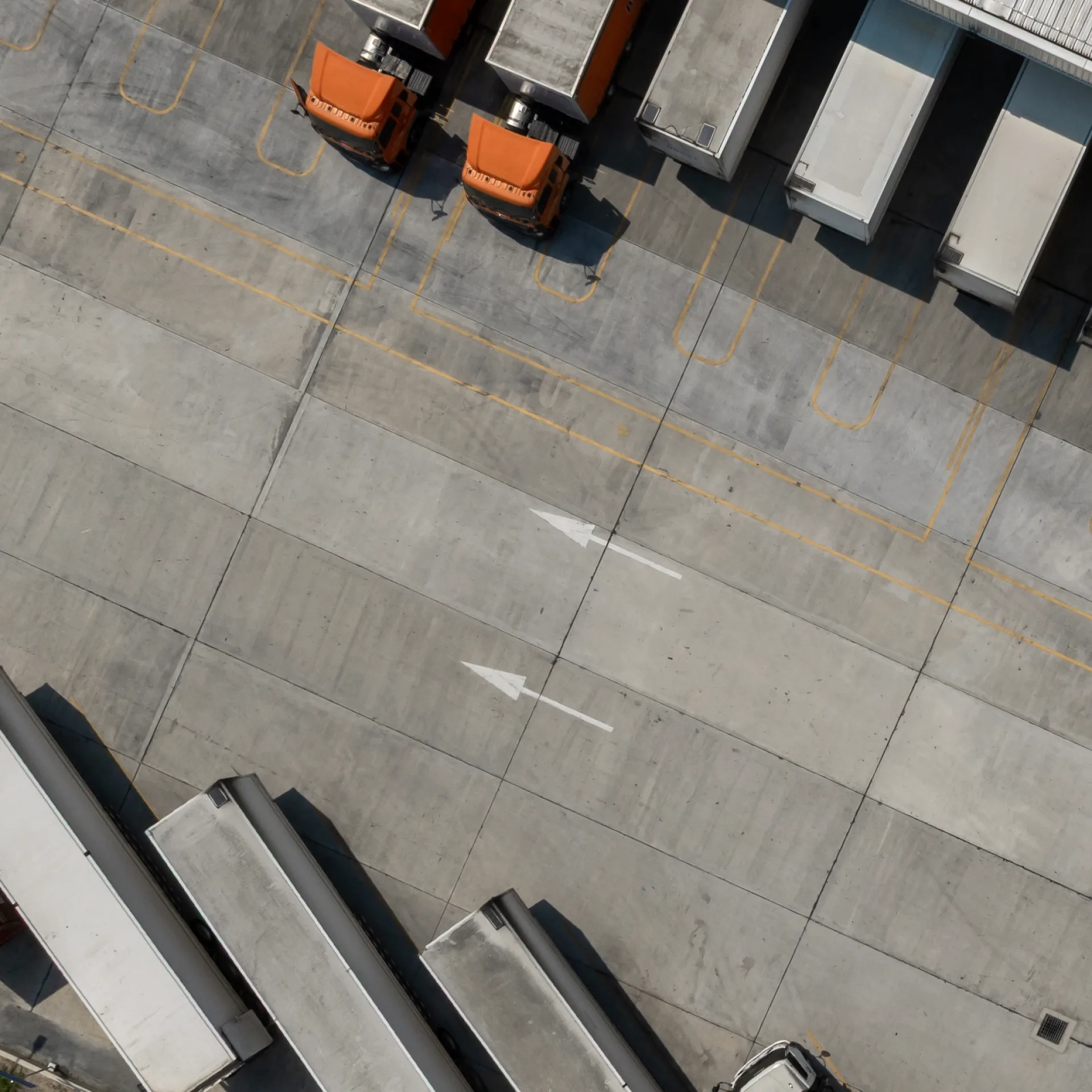 Aerial view of an industrial loading dock with three orange trucks parked at cargo bays. Concrete pavement shows marked lanes and arrows, conveying order.