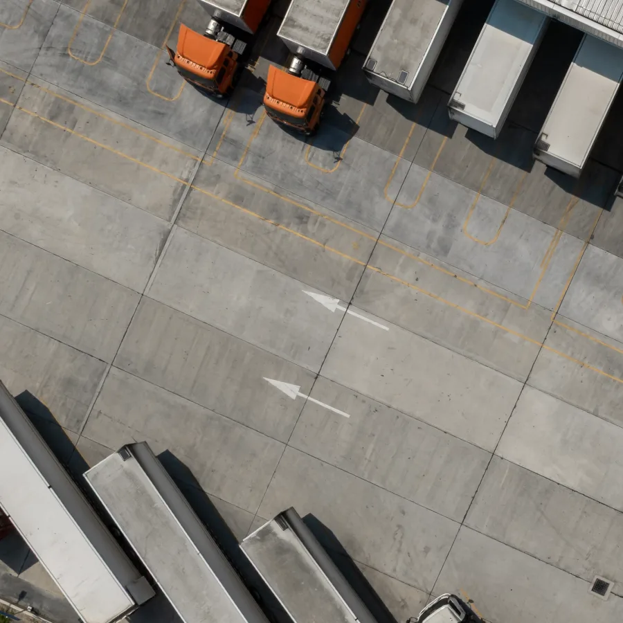 Aerial view of an industrial loading dock with three orange trucks parked at cargo bays. Concrete pavement shows marked lanes and arrows, conveying order.