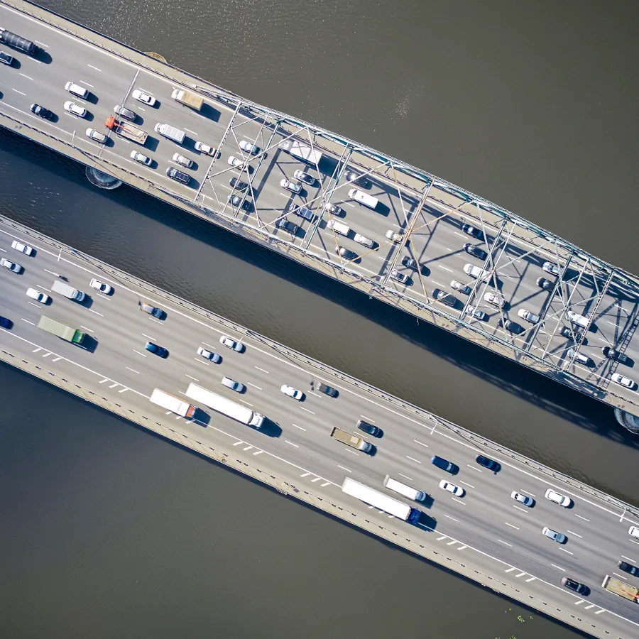 Aerial view of two parallel bridges over a river, filled with cars and trucks in heavy traffic. The scene conveys a sense of busyness and congestion.