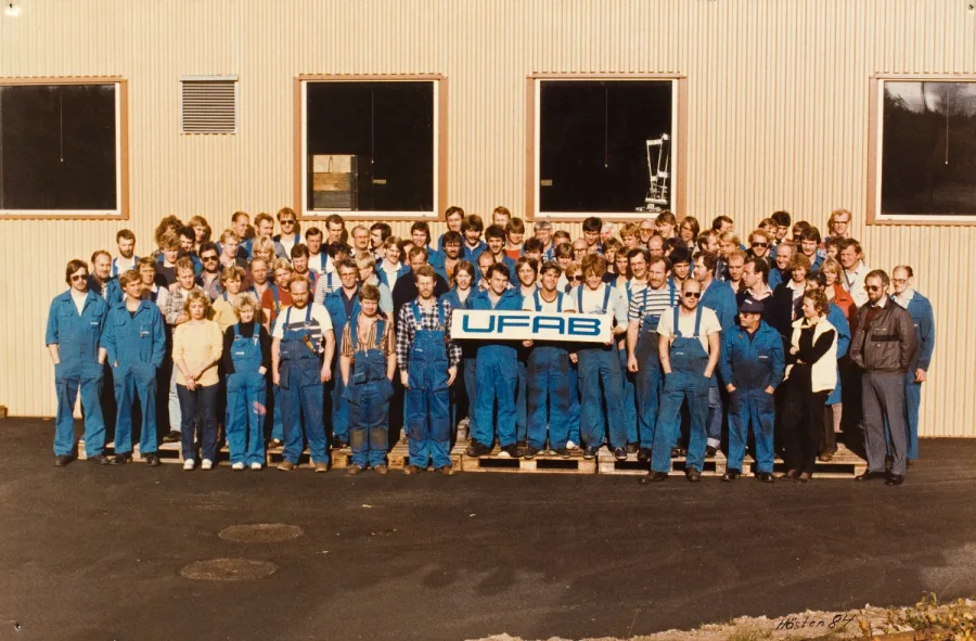 A large group of people, wearing blue coveralls, stand in rows outside a building holding a