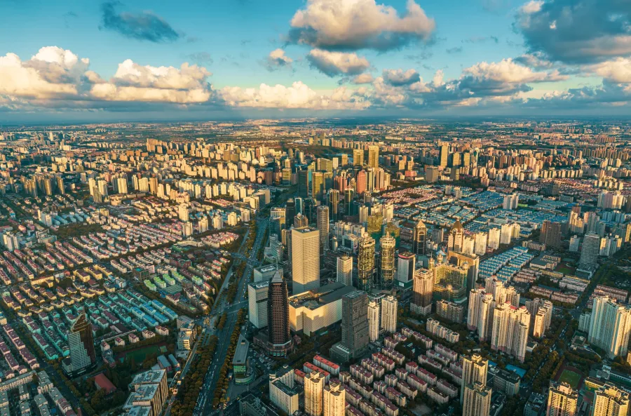 Aerial view of a sprawling cityscape at sunset with dense clusters of tall buildings, casting long shadows. Partly cloudy sky adds depth and drama.