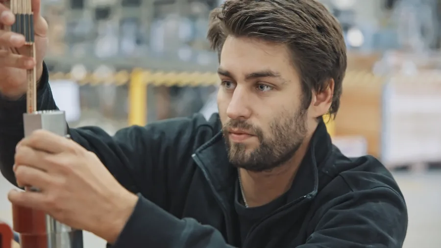 An image of a person working in a factory. The person is measuring a piece of metal.