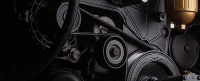 Close-up of a complex engine with black belts, gears, and metal components. A brass-colored filter stands out, conveying a technical and industrial feel.