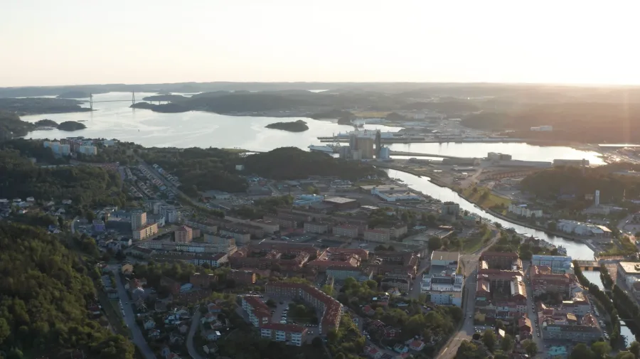 Aerial view of a coastal city at sunset. Buildings and roads are surrounded by lush green hills, with a river and bridges in the background. Calm atmosphere.